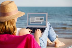 Rear view shot of a woman using her laptop and while sitting on beach. Work remotely