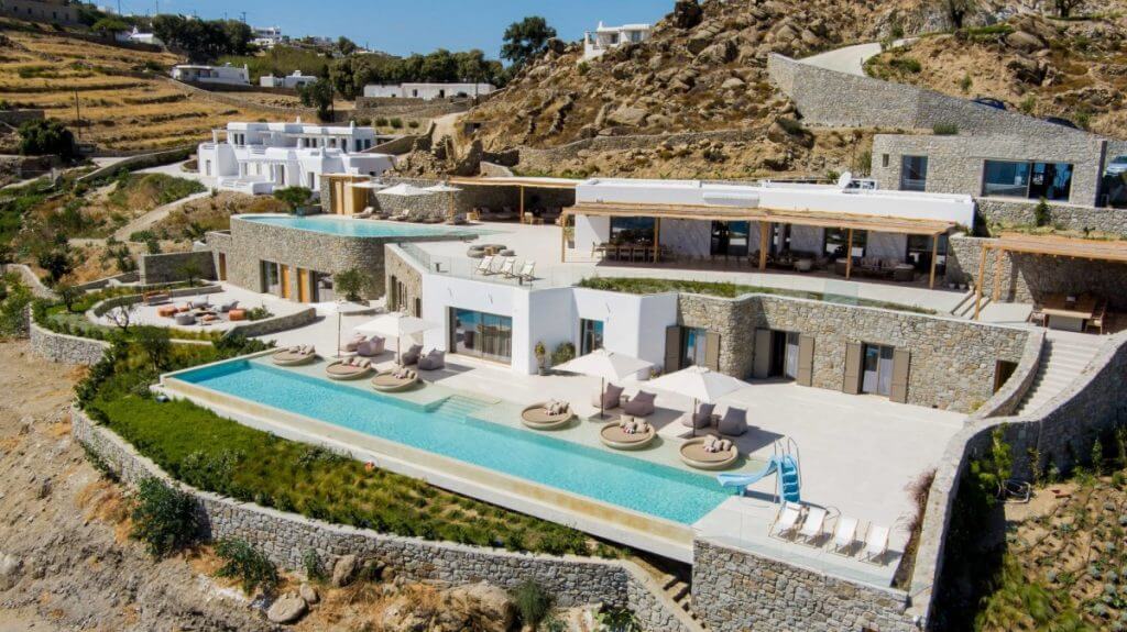 Luxury clifftop villa in Mykonos with panoramic pools, stone architecture, and sun loungers overlooking the Aegean landscape.