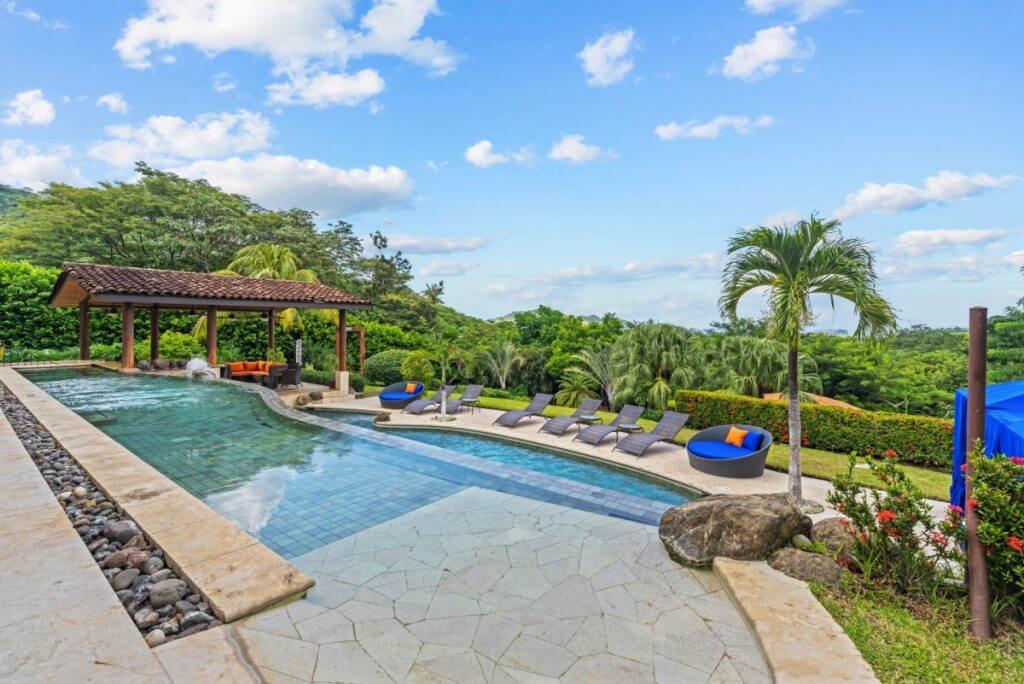 Villa Buena Onda, Costa Rica infinity pool with loungers, cabana, palm trees, and lush gardens under a clear sky.