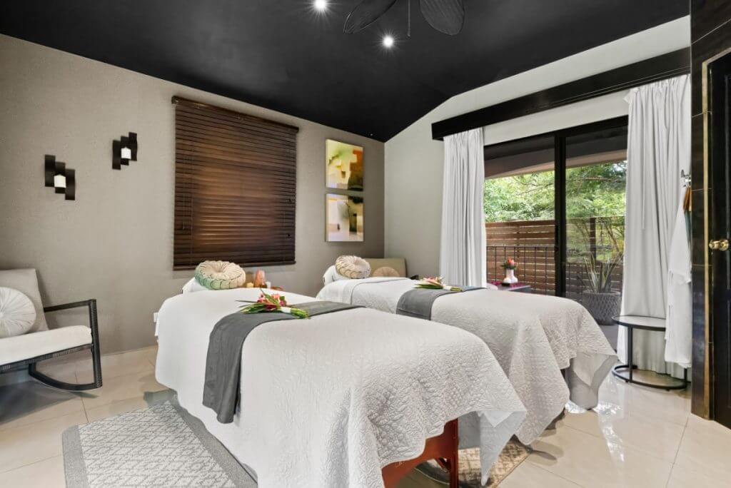 Spa room with two massage tables, serene decor, and tropical greenery outside.