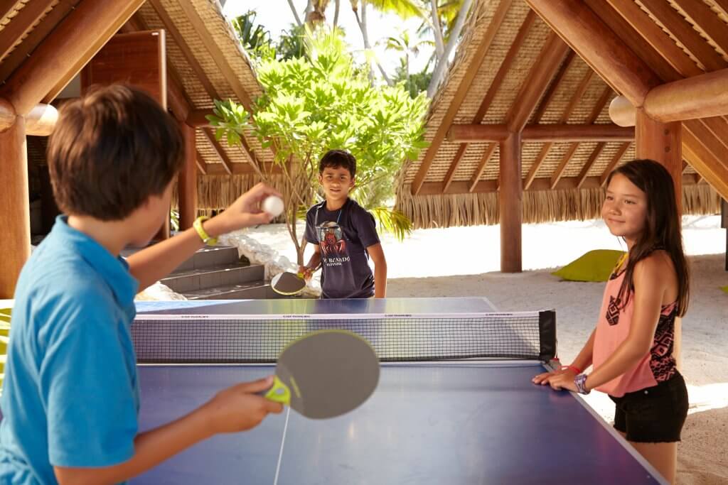 Kids playing table tennis in a shaded area.