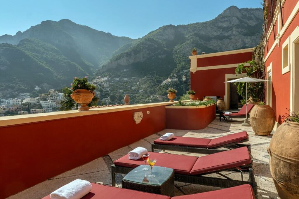 Terrace at Dorata villa on Amalfi Coast with red loungers, drinks, umbrella, and panoramic mountain views.