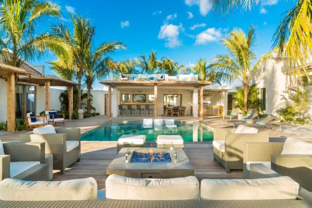 Ambergris Cay luxury villa with pool, sunken loungers, firepit seating, palm trees, and open-air bar under clear tropical skies.